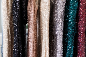 A series of sequin-covered dresses hanging on a rack, displaying a variety of colors including black, beige, gold, silver, teal, and dark red. The sequins create a shimmering effect as they catch the light.