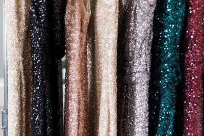 A series of sequin-covered dresses hanging on a rack, displaying a variety of colors including black, beige, gold, silver, teal, and dark red. The sequins create a shimmering effect as they catch the light.