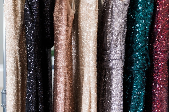 A series of sequin-covered dresses hanging on a rack, displaying a variety of colors including black, beige, gold, silver, teal, and dark red. The sequins create a shimmering effect as they catch the light.