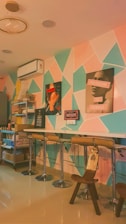 An interior setting featuring a modern bar area with high stools and a counter against a geometric-patterned wall. There are various artworks displayed, including images of a woman and slogans. Lighting is warm and enhances the cozy atmosphere.