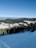 A scenic view of a ski resort nestled in a mountainous region. There is a wide expanse of snow-covered slopes in the foreground, surrounded by dense evergreen forests. In the background, several resort buildings with distinct architecture are visible, set against the backdrop of rolling hills and a clear blue sky.