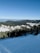 A scenic view of a ski resort nestled in a mountainous region. There is a wide expanse of snow-covered slopes in the foreground, surrounded by dense evergreen forests. In the background, several resort buildings with distinct architecture are visible, set against the backdrop of rolling hills and a clear blue sky.