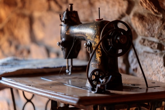 An antique sewing machine with intricate gold detailing stands on a wooden table. The ambiance is warm with a blurred stone wall backdrop, creating a rustic atmosphere.