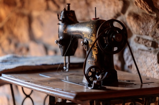 An antique sewing machine with intricate gold detailing stands on a wooden table. The ambiance is warm with a blurred stone wall backdrop, creating a rustic atmosphere.