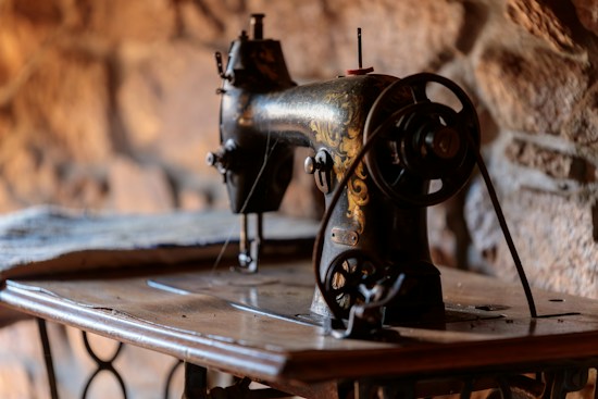 An antique sewing machine with intricate gold detailing stands on a wooden table. The ambiance is warm with a blurred stone wall backdrop, creating a rustic atmosphere.