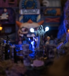 A mysterious figure clad in a dark hooded robe holds two glowing orbs, surrounded by a dimly lit, eerie setting with bluish lights and a spooky background featuring unusual structures.