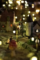 A miniature scene depicting a quaint, picturesque village at night. The setting includes small houses with textured roofs and lit windows, surrounded by tiny trees and illuminated by delicate streetlamps. The soft glow of the lights creates a cozy and enchanting atmosphere, enhancing the charm of the village.