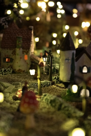 A miniature scene depicting a quaint, picturesque village at night. The setting includes small houses with textured roofs and lit windows, surrounded by tiny trees and illuminated by delicate streetlamps. The soft glow of the lights creates a cozy and enchanting atmosphere, enhancing the charm of the village.