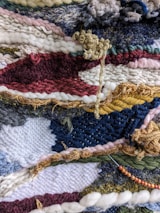A close-up view of a colorful, textured woven textile with a variety of yarns and fibers intricately woven together. The pattern features stripes and abstract shapes with different weaving techniques, creating a dynamic and intricate surface.