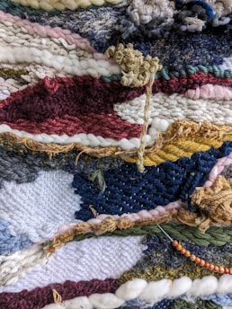 A close-up view of a colorful, textured woven textile with a variety of yarns and fibers intricately woven together. The pattern features stripes and abstract shapes with different weaving techniques, creating a dynamic and intricate surface.