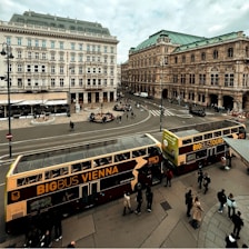 A bustling urban scene featuring a historic European square with grand architectural buildings. Two red and yellow double-decker buses labeled 'BIG BUS VIENNA' are in the foreground, with several pedestrians walking around and a few people sitting at a circular seating area. The sky is overcast, and the square is surrounded by ornate, historic buildings.