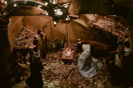 A nativity scene featuring figurines of Mary, Joseph, and baby Jesus in a manger surrounded by animals like oxen, with straw scattered around. A star adorned with lights hangs above, casting a warm glow over the setting.