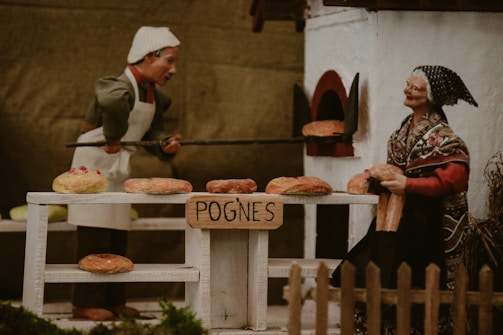 Two figures are depicted in a bakery scene with one baking bread in an oven and the other standing nearby. Several loaves of bread are displayed on a table labeled 'POGNES'.