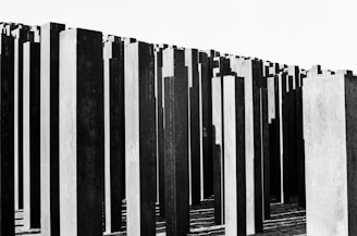 Numerous tall, narrow concrete slabs are arranged in a grid-like pattern, creating a vast and somber architectural structure. The slabs vary in height and are positioned closely together, casting deep shadows on the ground.