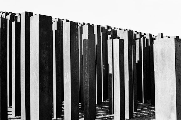 Numerous tall, narrow concrete slabs are arranged in a grid-like pattern, creating a vast and somber architectural structure. The slabs vary in height and are positioned closely together, casting deep shadows on the ground.