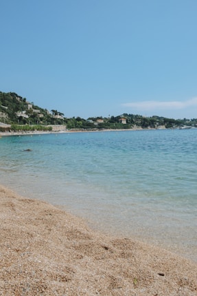 A serene beach with golden sand gently slopes into clear blue water. In the background, lush green hills are dotted with houses. The sky is clear and blue, adding to the tranquil atmosphere.