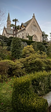 An old stone church with a triangular roof and a small spire is surrounded by lush greenery, including bushes and trees. The foreground features well-maintained hedges and grass, contributing to a serene garden setting. Palm trees are visible alongside the church, adding an exotic touch to the scene.