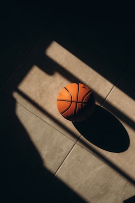 A basketball sits on a tiled floor, partially illuminated by light coming through a window. The shadows create a dramatic effect, highlighting the patterns on the ball and the texture of the tiles.