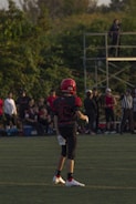 A young football player stands on a grassy field, wearing a red helmet and black uniform with the number 36. Other players and spectators are visible in the background, with trees and a metal scaffold structure nearby.