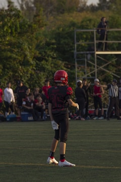 A young football player stands on a grassy field, wearing a red helmet and black uniform with the number 36. Other players and spectators are visible in the background, with trees and a metal scaffold structure nearby.