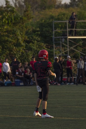 A young football player stands on a grassy field, wearing a red helmet and black uniform with the number 36. Other players and spectators are visible in the background, with trees and a metal scaffold structure nearby.