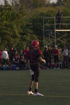A young football player stands on a grassy field, wearing a red helmet and black uniform with the number 36. Other players and spectators are visible in the background, with trees and a metal scaffold structure nearby.