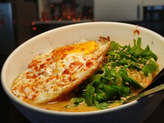 A bowl of ramen with a rich, creamy broth featuring a perfectly fried egg on top. The dish is garnished with fresh green onions, sesame seeds, and chili flakes. The noodles and various toppings are slightly submerged, creating a visually appealing combination of textures and colors.