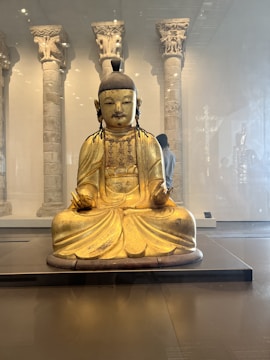 A golden Buddha statue is seated in a lotus position in a museum setting, with intricately carved stone pillars in the background. The statue has delicate features and is adorned with simple jewelry, and the room is softly lit.