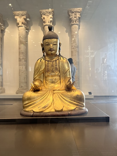 A golden Buddha statue is seated in a lotus position in a museum setting, with intricately carved stone pillars in the background. The statue has delicate features and is adorned with simple jewelry, and the room is softly lit.