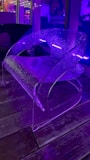 A translucent modern chair with a sleek, curved design is illuminated by purple lighting. The setting appears to be indoors with reflections of additional light sources visible in the background. The floor is wooden, adding a contrast to the contemporary feel of the chair.