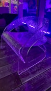 A translucent modern chair with a sleek, curved design is illuminated by purple lighting. The setting appears to be indoors with reflections of additional light sources visible in the background. The floor is wooden, adding a contrast to the contemporary feel of the chair.
