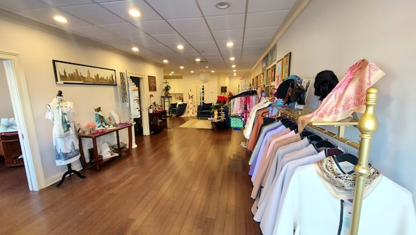 The image depicts the interior of a boutique with neatly arranged clothing on racks along the right side. The floor is made of polished wood, and the walls are adorned with artwork and framed pictures. There are mannequins dressed in various outfits and displays of accessories on tables. The overall setup is elegant and organized.