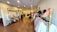 The image depicts the interior of a boutique with neatly arranged clothing on racks along the right side. The floor is made of polished wood, and the walls are adorned with artwork and framed pictures. There are mannequins dressed in various outfits and displays of accessories on tables. The overall setup is elegant and organized.