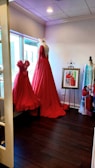 A room featuring two elegant red dresses displayed on mannequins near a large window. The floor-length gown has a deep back and intricate detailing. A framed picture of a similar dress stands on an easel, alongside another mannequin draped with a colorful fabric. The room has a warm ambiance accentuated by the wooden floor and soft lighting.