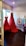 A room featuring two elegant red dresses displayed on mannequins near a large window. The floor-length gown has a deep back and intricate detailing. A framed picture of a similar dress stands on an easel, alongside another mannequin draped with a colorful fabric. The room has a warm ambiance accentuated by the wooden floor and soft lighting.