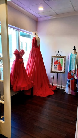 A room featuring two elegant red dresses displayed on mannequins near a large window. The floor-length gown has a deep back and intricate detailing. A framed picture of a similar dress stands on an easel, alongside another mannequin draped with a colorful fabric. The room has a warm ambiance accentuated by the wooden floor and soft lighting.