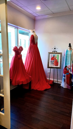 A room featuring two elegant red dresses displayed on mannequins near a large window. The floor-length gown has a deep back and intricate detailing. A framed picture of a similar dress stands on an easel, alongside another mannequin draped with a colorful fabric. The room has a warm ambiance accentuated by the wooden floor and soft lighting.