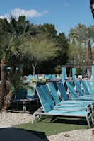 A series of turquoise lounge chairs are arranged neatly by a poolside, accompanied by a backdrop of lush palm trees and greenery. The area is well-maintained with stone pathways and patches of artificial grass. A section of the pool is visible surrounded by relaxing daybeds and shaded cabanas.
