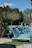 A series of turquoise lounge chairs are arranged neatly by a poolside, accompanied by a backdrop of lush palm trees and greenery. The area is well-maintained with stone pathways and patches of artificial grass. A section of the pool is visible surrounded by relaxing daybeds and shaded cabanas.