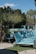 A series of turquoise lounge chairs are arranged neatly by a poolside, accompanied by a backdrop of lush palm trees and greenery. The area is well-maintained with stone pathways and patches of artificial grass. A section of the pool is visible surrounded by relaxing daybeds and shaded cabanas.