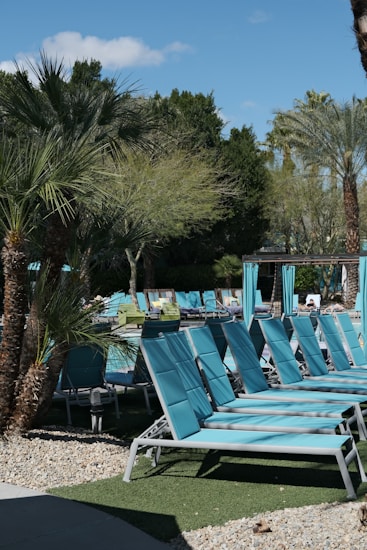 A series of turquoise lounge chairs are arranged neatly by a poolside, accompanied by a backdrop of lush palm trees and greenery. The area is well-maintained with stone pathways and patches of artificial grass. A section of the pool is visible surrounded by relaxing daybeds and shaded cabanas.