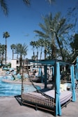 This image features a resort-like outdoor pool area. There are several cabanas with blue drapes and lounge chairs situated closely around the swimming pool. Palm trees are scattered throughout the scene, giving a tropical vibe. People can be seen lounging and enjoying the sunny weather. The sky is clear and blue.