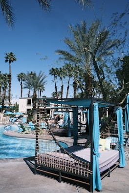 This image features a resort-like outdoor pool area. There are several cabanas with blue drapes and lounge chairs situated closely around the swimming pool. Palm trees are scattered throughout the scene, giving a tropical vibe. People can be seen lounging and enjoying the sunny weather. The sky is clear and blue.