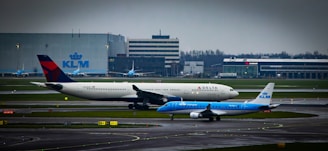 Two airplanes, one from KLM and another from Delta, are positioned on a runway at an airport. In the background, there are airport buildings and hangars, including a prominent KLM building. The sky is overcast, enhancing the industrial feel of the setting.