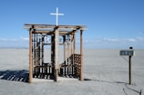 A rustic outdoor confessional booth sits alone in a barren desert landscape under a clear blue sky. The structure is made of wood with a cross on top and has the word 'confession' painted on it. A solitary mailbox, adorned with small crosses, stands to the right.