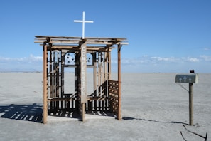 A rustic outdoor confessional booth sits alone in a barren desert landscape under a clear blue sky. The structure is made of wood with a cross on top and has the word 'confession' painted on it. A solitary mailbox, adorned with small crosses, stands to the right.