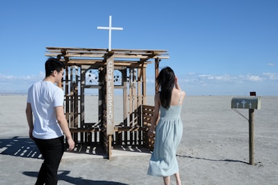 A wooden structure with the word 'Confession' written on it stands in a deserted, arid landscape. Two people, a man wearing a white t-shirt and black pants and a woman in a light blue dress, are walking towards the structure. A small mailbox with a cross design is positioned nearby. The sky is clear with a few clouds visible in the distance.