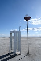 An old-fashioned phone booth labeled 'FREE LOVE' stands alone on a barren, flat expanse. Next to it is a tall pole with a large red heart near the top, connected to the booth by a wire. The sky is clear with scattered clouds.