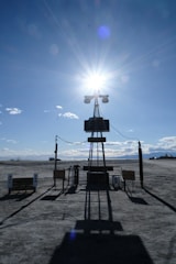 A barren landscape with a large solar panel structure in the foreground, casting a long shadow on the ground. The sun shines brightly behind it, creating a starburst effect in a clear blue sky. Sparse clouds dot the horizon, and there is a vehicle parked in the distance.