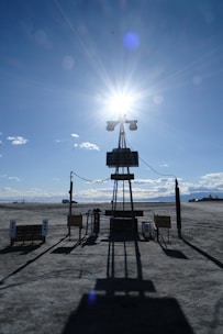 A barren landscape with a large solar panel structure in the foreground, casting a long shadow on the ground. The sun shines brightly behind it, creating a starburst effect in a clear blue sky. Sparse clouds dot the horizon, and there is a vehicle parked in the distance.